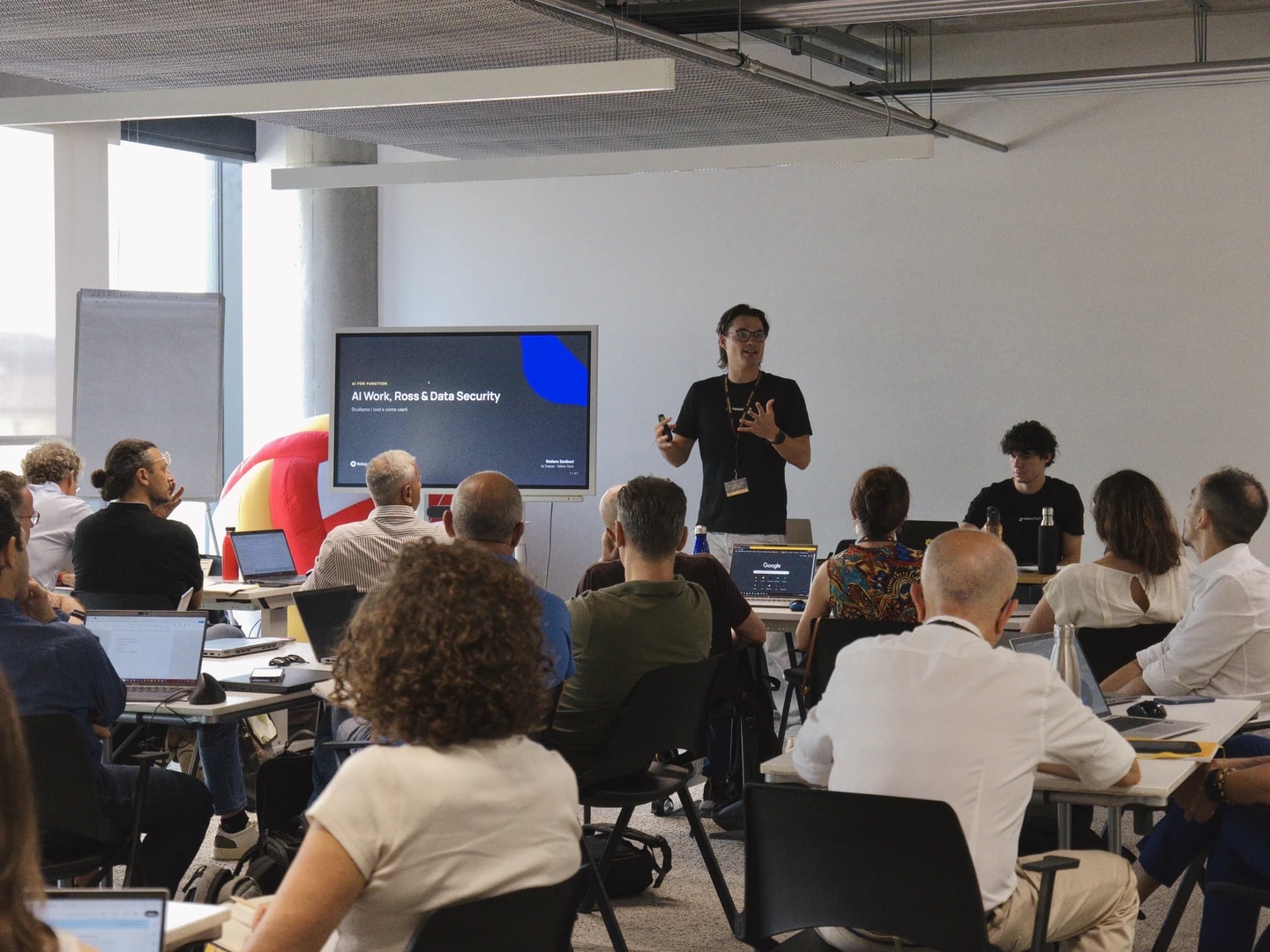 Un formatore Yellow Tech parla davanti a un'aula piena di partecipanti seduti ai tavoli con i portatili, accanto allo schermo della presentazione